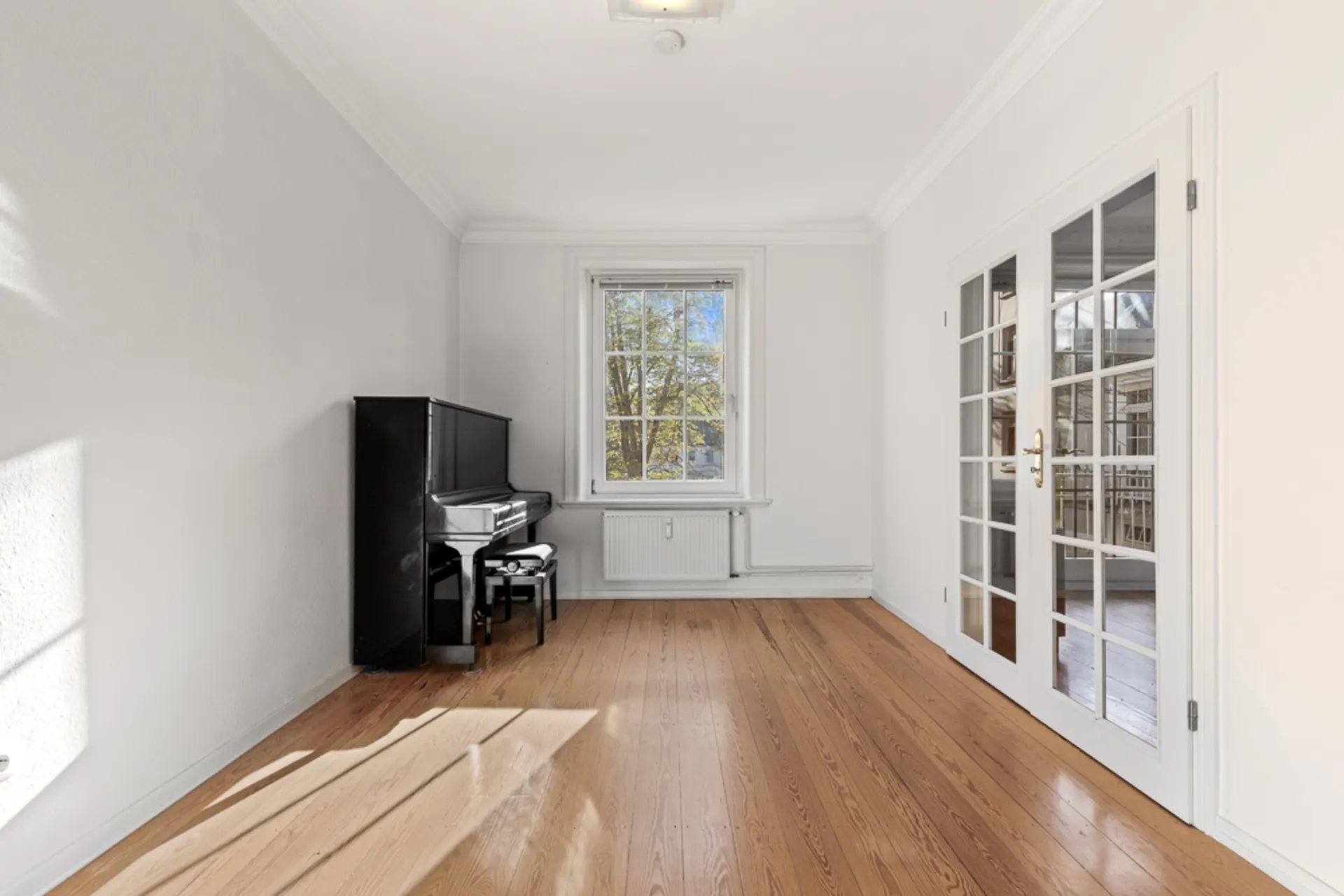 Helles Zimmer mit Holzboden, Fenster, Glastüren und schwarzem Klavier.