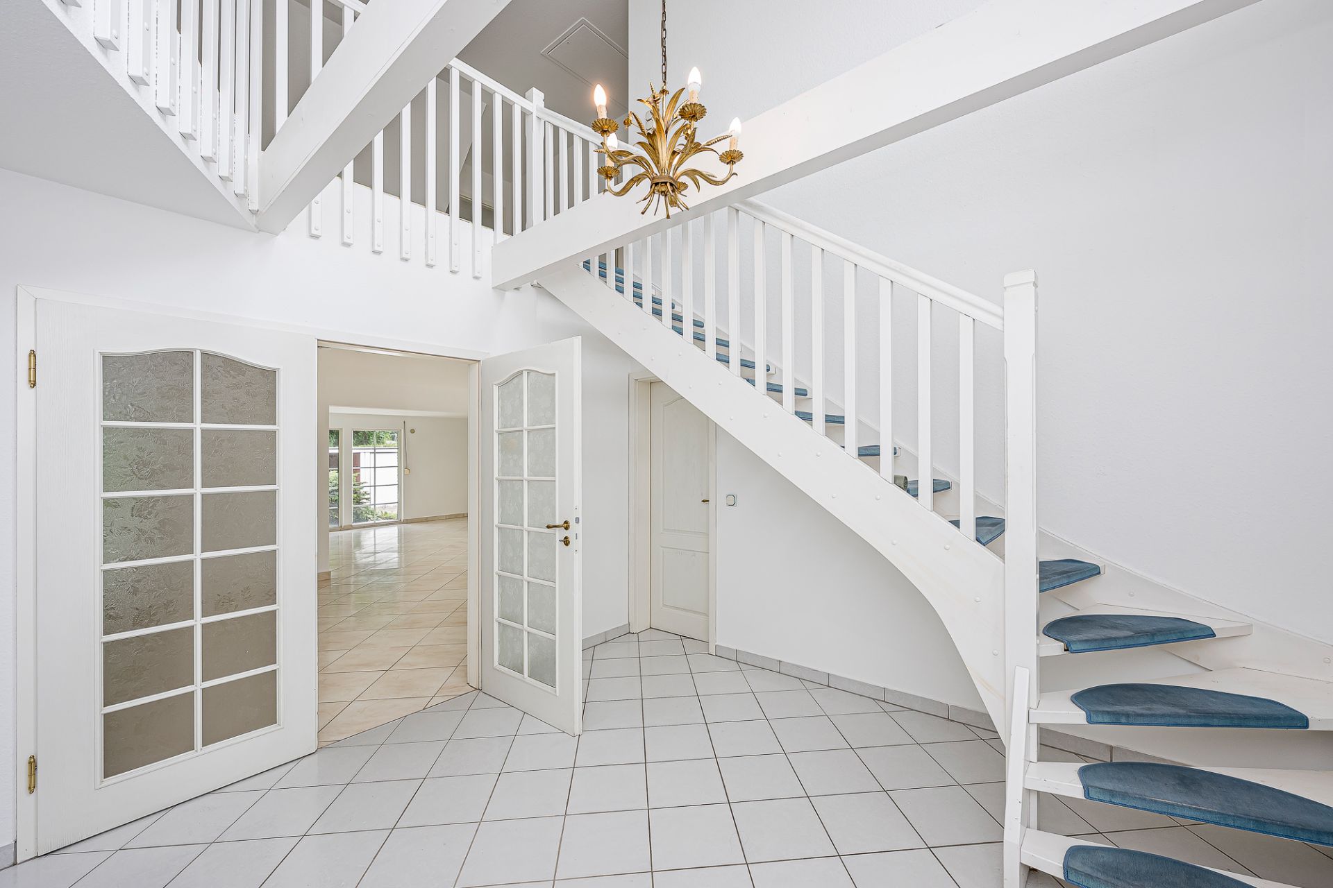 Heller Flur mit weißer Treppe, Kronleuchter und Flügeltüren zu einem großen Raum.