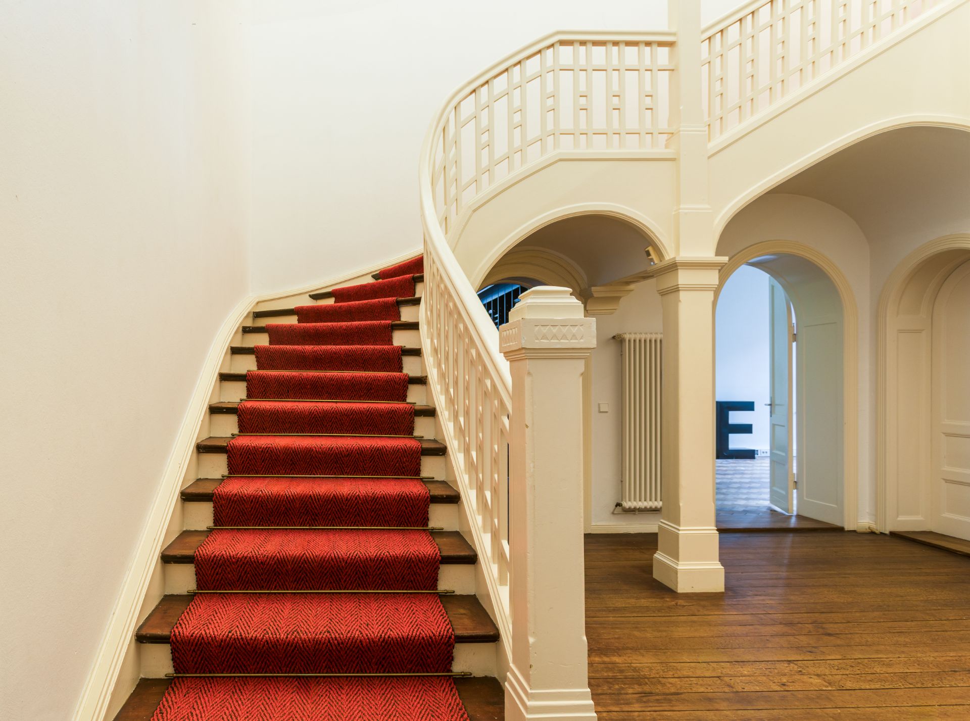 Geschwungene Treppe mit rotem Teppich und weißem Geländer in einem Flur mit Holzboden.