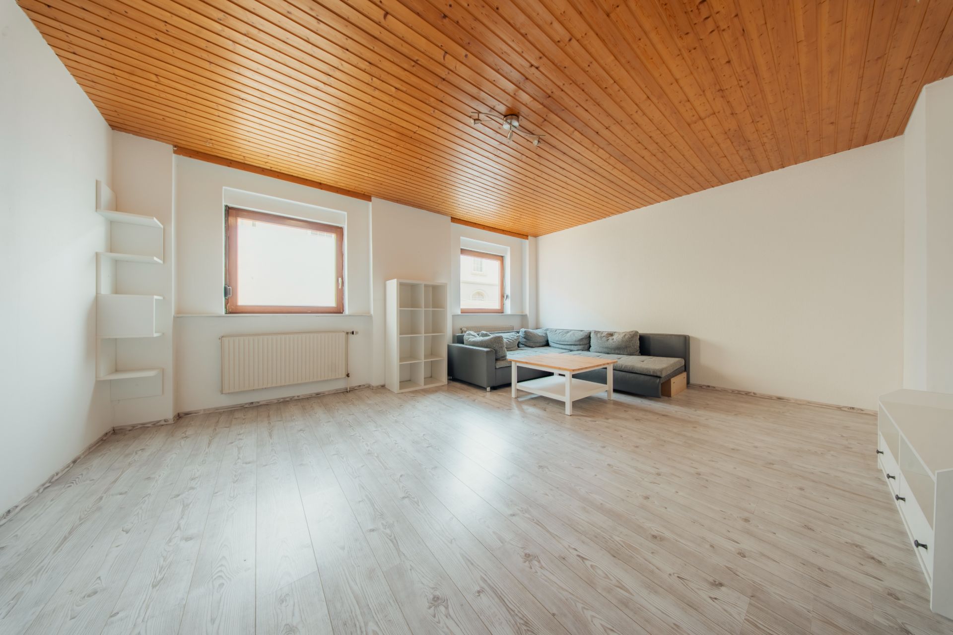 Helles Wohnzimmer mit Holzdecke, Ecksofa, weißem Tisch und Regal, zwei Fenstern und Wandregalen.