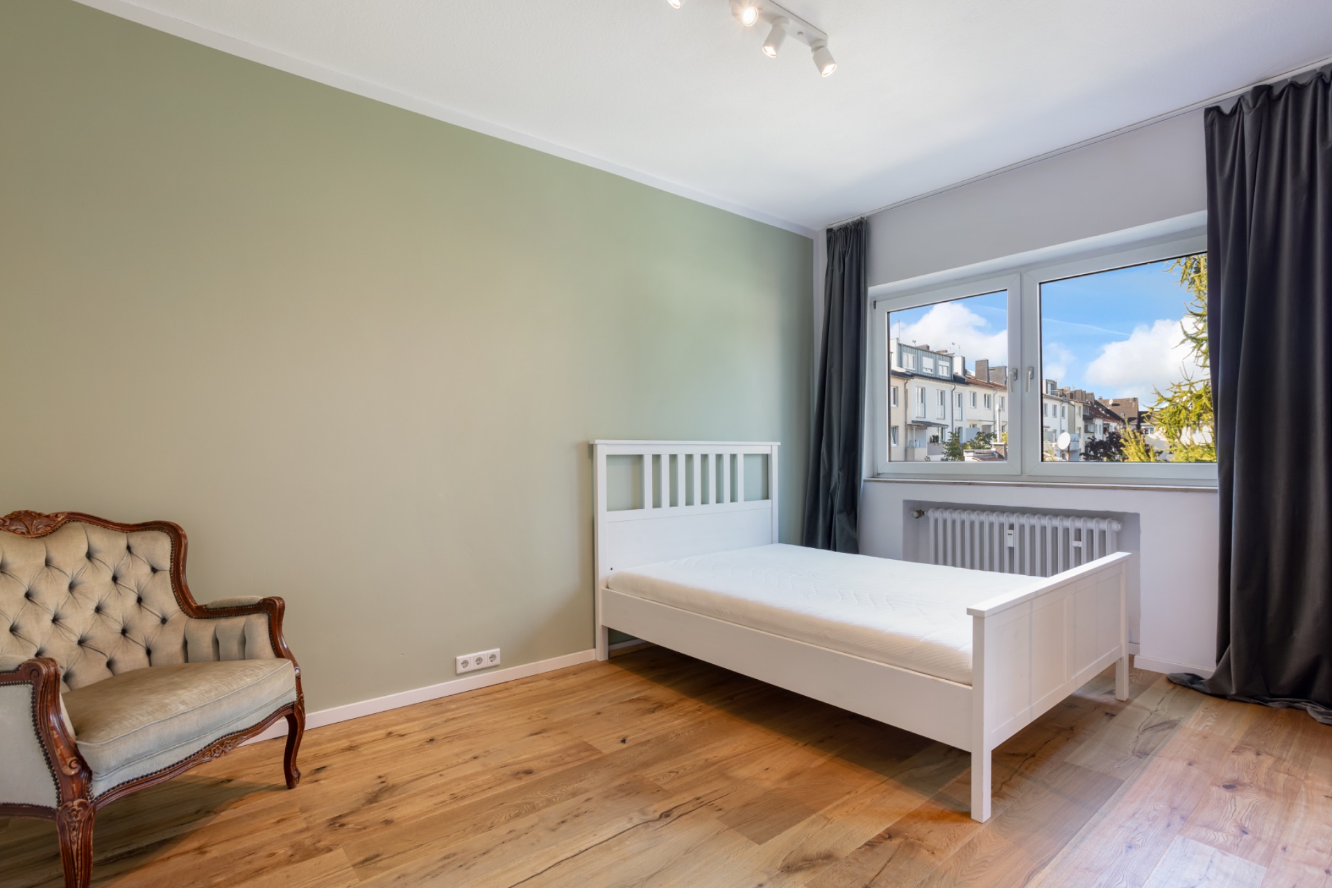 Helles Schlafzimmer mit weißem Bett, Sessel, Holzfußboden und großem Fenster mit Vorhängen.