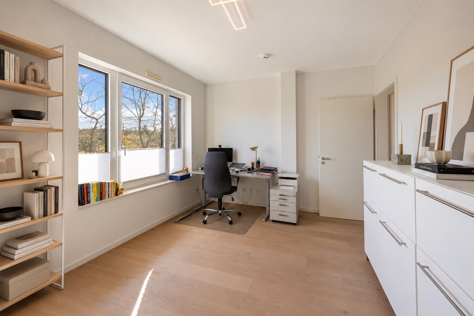 Helles Arbeitszimmer mit Schreibtisch, Bürostuhl, Regal und großem Fenster.