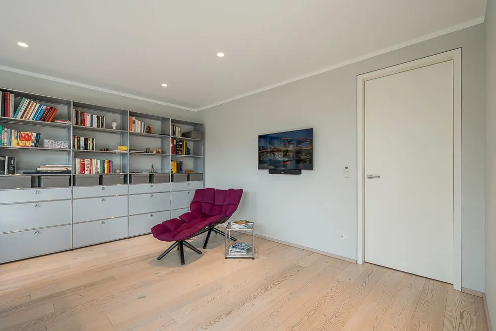 Modernes Zimmer mit Bücherregal, rotem Sessel, Wand-TV und hellen Holzboden.