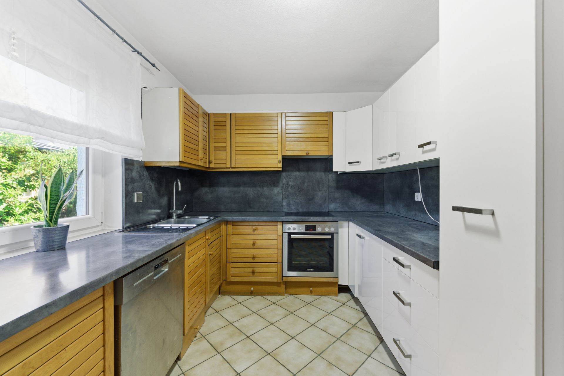 Moderne Küche mit Holzelementen, weißen Schränken, Backofen, Spüle und großem Fenster mit Pflanze.