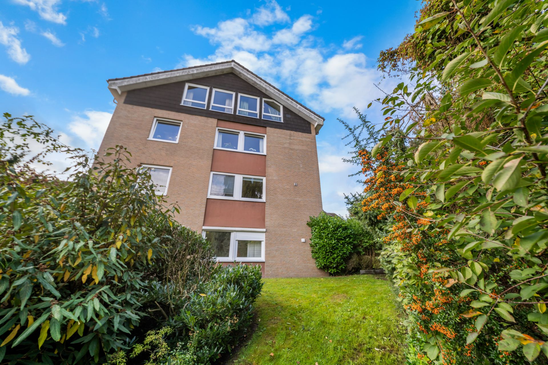 Mehrfamilienhaus mit vier Stockwerken, umgeben von Garten und Büschen, unter blauem Himmel.