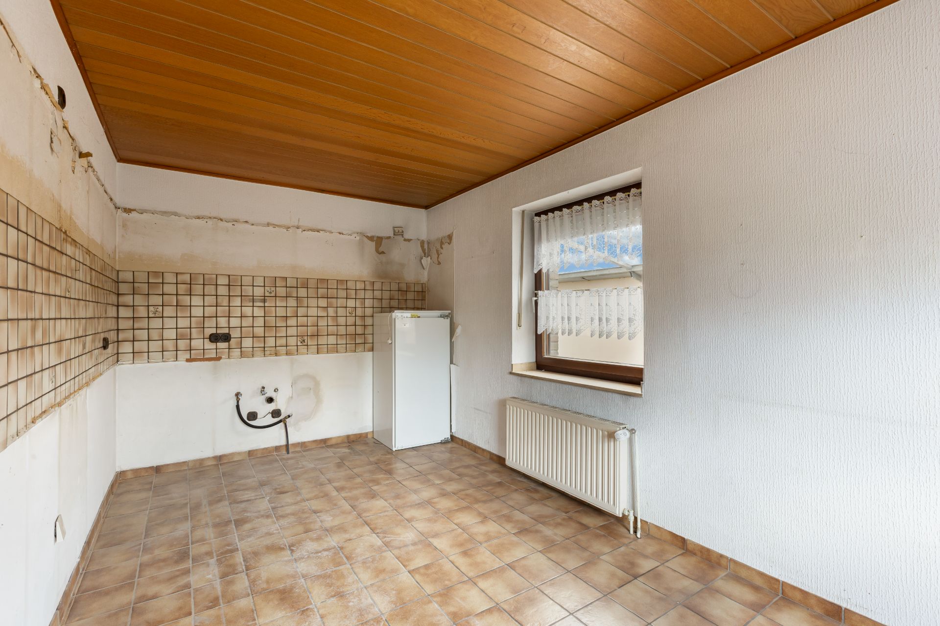 Leerer Raum mit Fliesenboden, Holzbalkendecke, Fenster, Heizkörper und kleinem Kühlschrank.