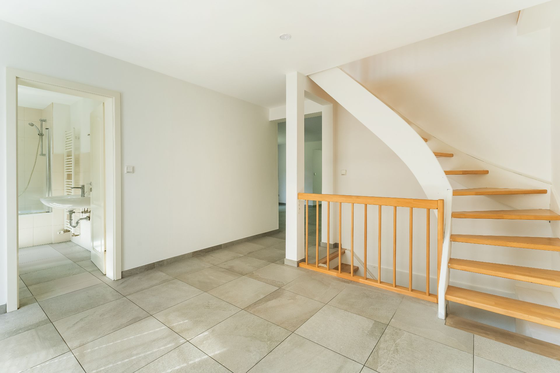 Helle Diele mit Fliesenboden, offener Holztreppe und Blick ins Badezimmer.