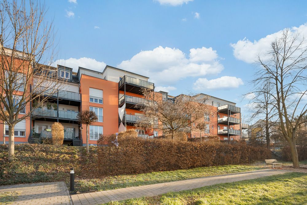 Mehrfamilienhaus mit Balkonen, Gehweg und Bäumen im Vordergrund bei sonnigem Wetter.