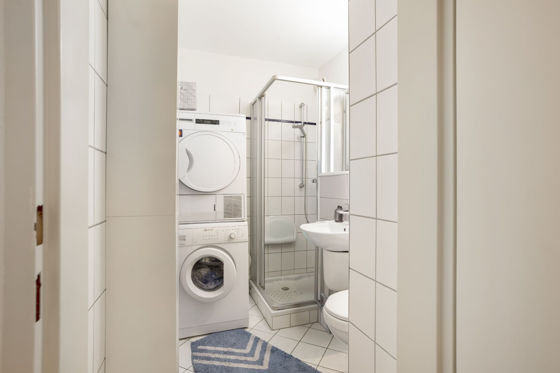 Kleines Bad mit Dusche, Waschmaschine, Trockner, Waschbecken, Toilette und blauem Teppich.