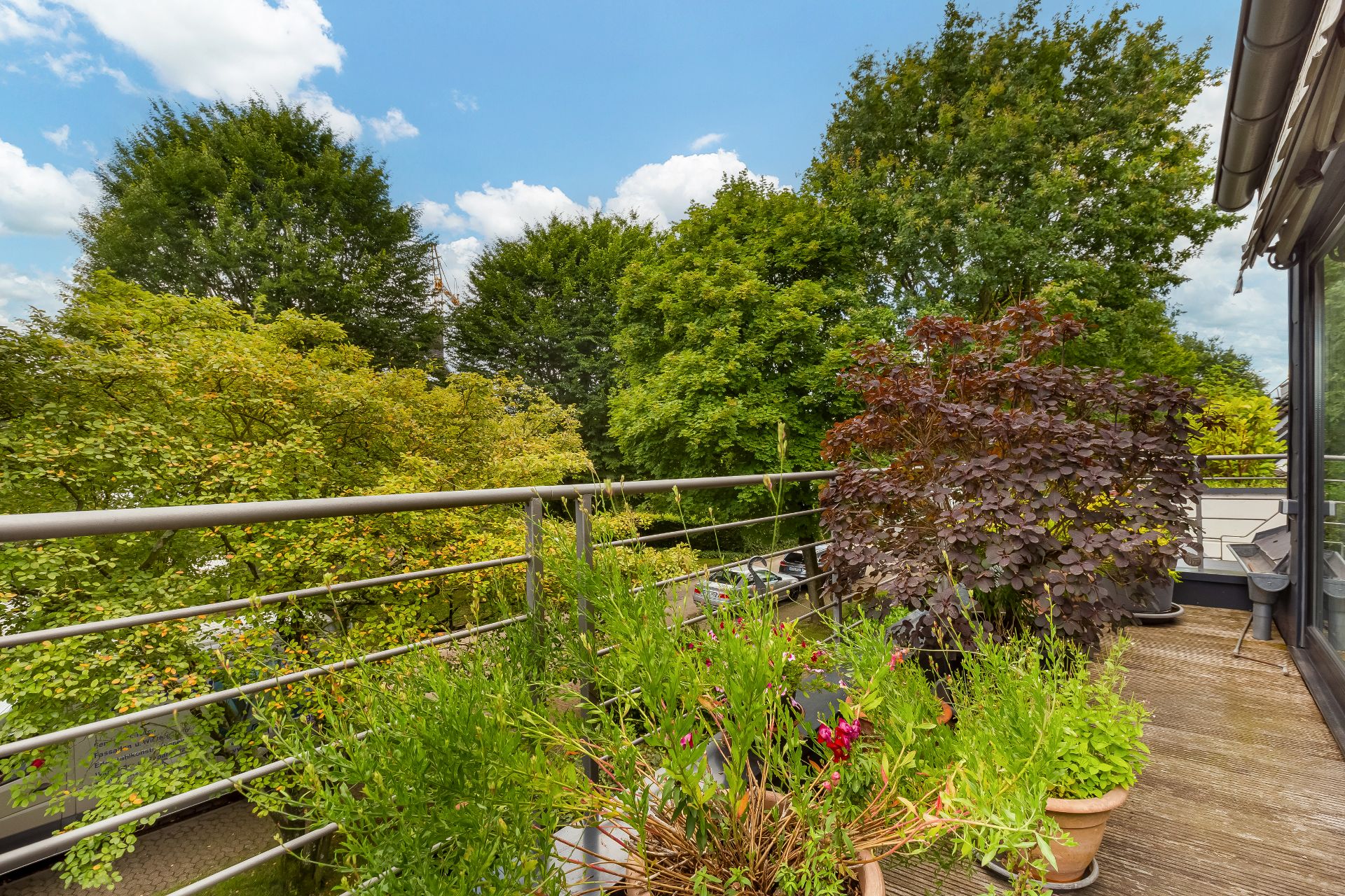 Balkon mit Blumenkästen und Pflanzen, umgeben von grünen Bäumen bei blauem Himmel.