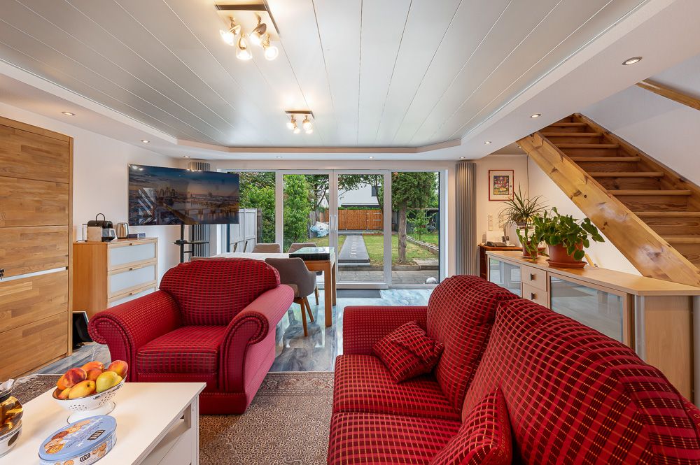 Helles Wohnzimmer mit roten Sofas, TV, Esstisch, Pflanzen und Blick in den Garten.
