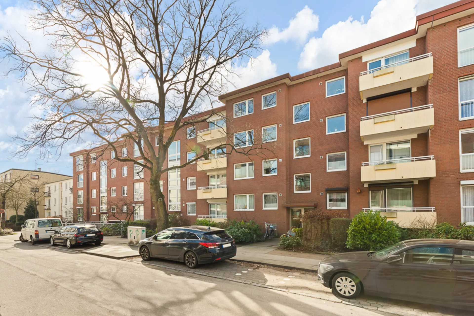 Mehrfamilienhaus aus rotem Backstein mit Balkonen, parkenden Autos und einem großen Baum davor.