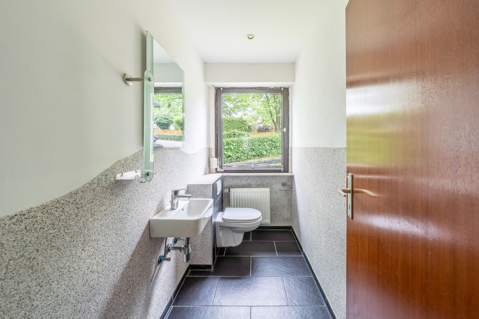 Helles Bad mit Fenster, Wandwaschbecken, Spiegel und Toilette auf dunklen Fliesen.