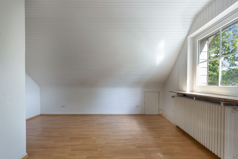 Leerer Dachbodenraum mit Holzfußboden, weißer Schräge und großem Fenster mit Heizung.