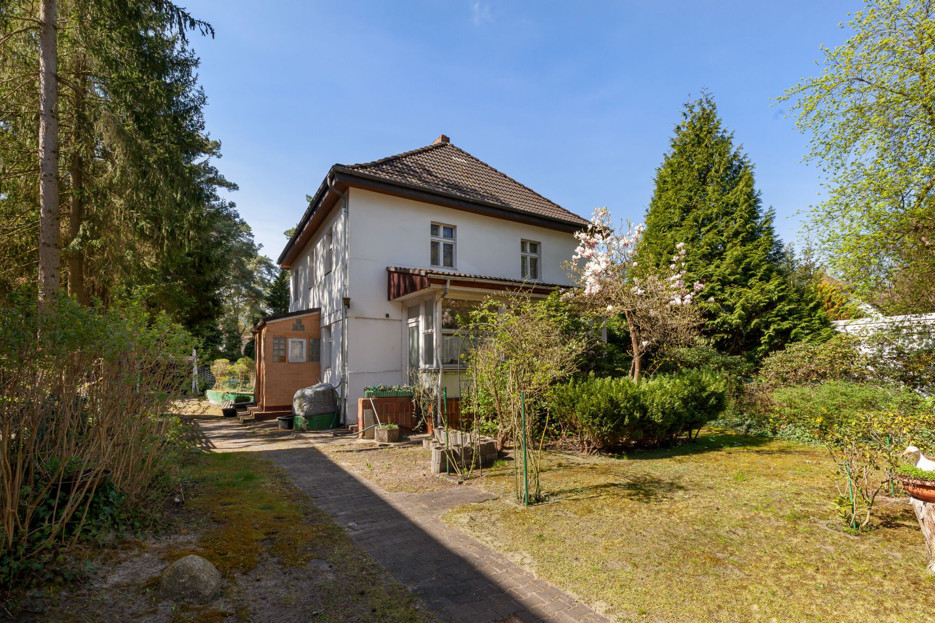 Zweistöckiges Haus mit Garten, umgeben von Bäumen und Sträuchern bei sonnigem Wetter.