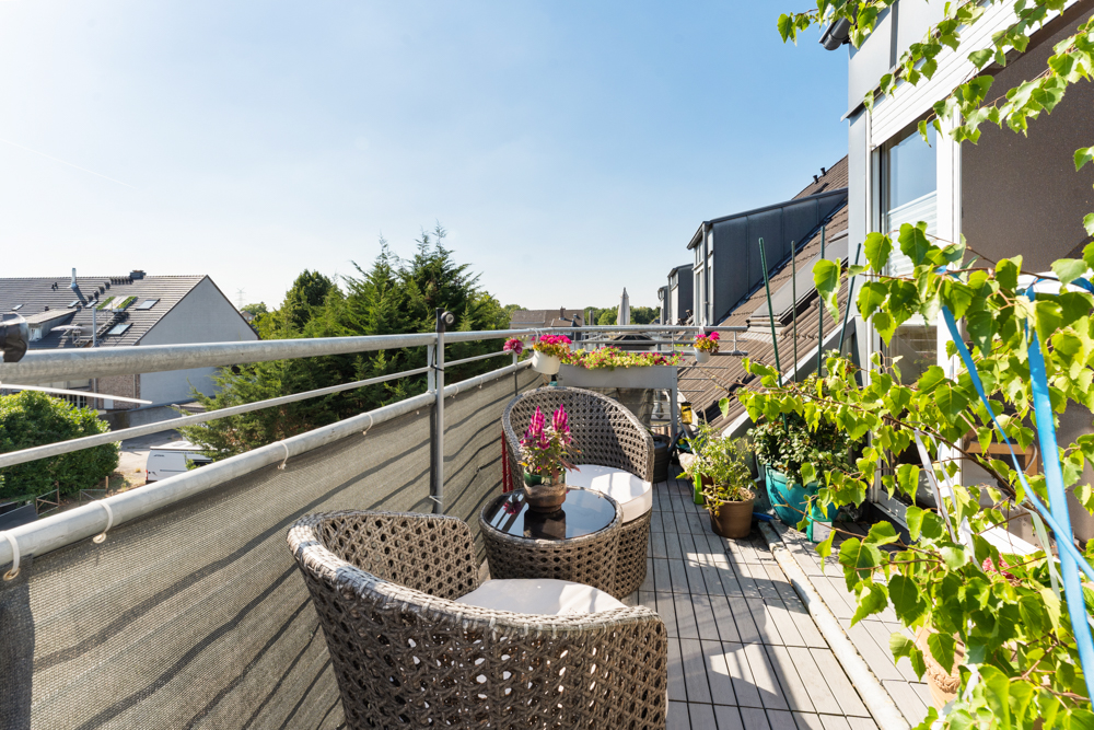 Balkon mit zwei Korbstühlen, kleinem Tisch und vielen Pflanzen bei sonnigem Wetter.