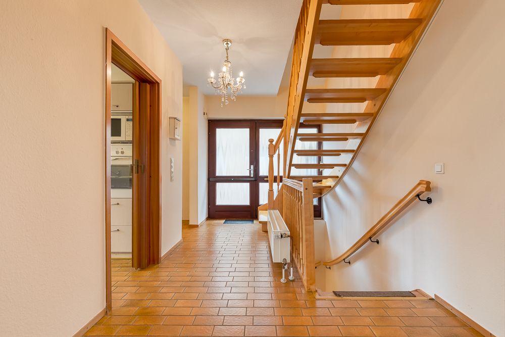 Heller Flur mit Holztreppe, Kronleuchter und braunen Fliesen, Tür zum Küchenbereich offen.