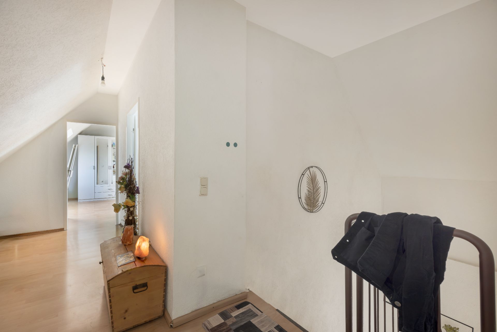 Heller Flur mit Holztruhe, Wanddeko und Handlauf, Boden aus Holz, Tür führt zu weiterem Raum.