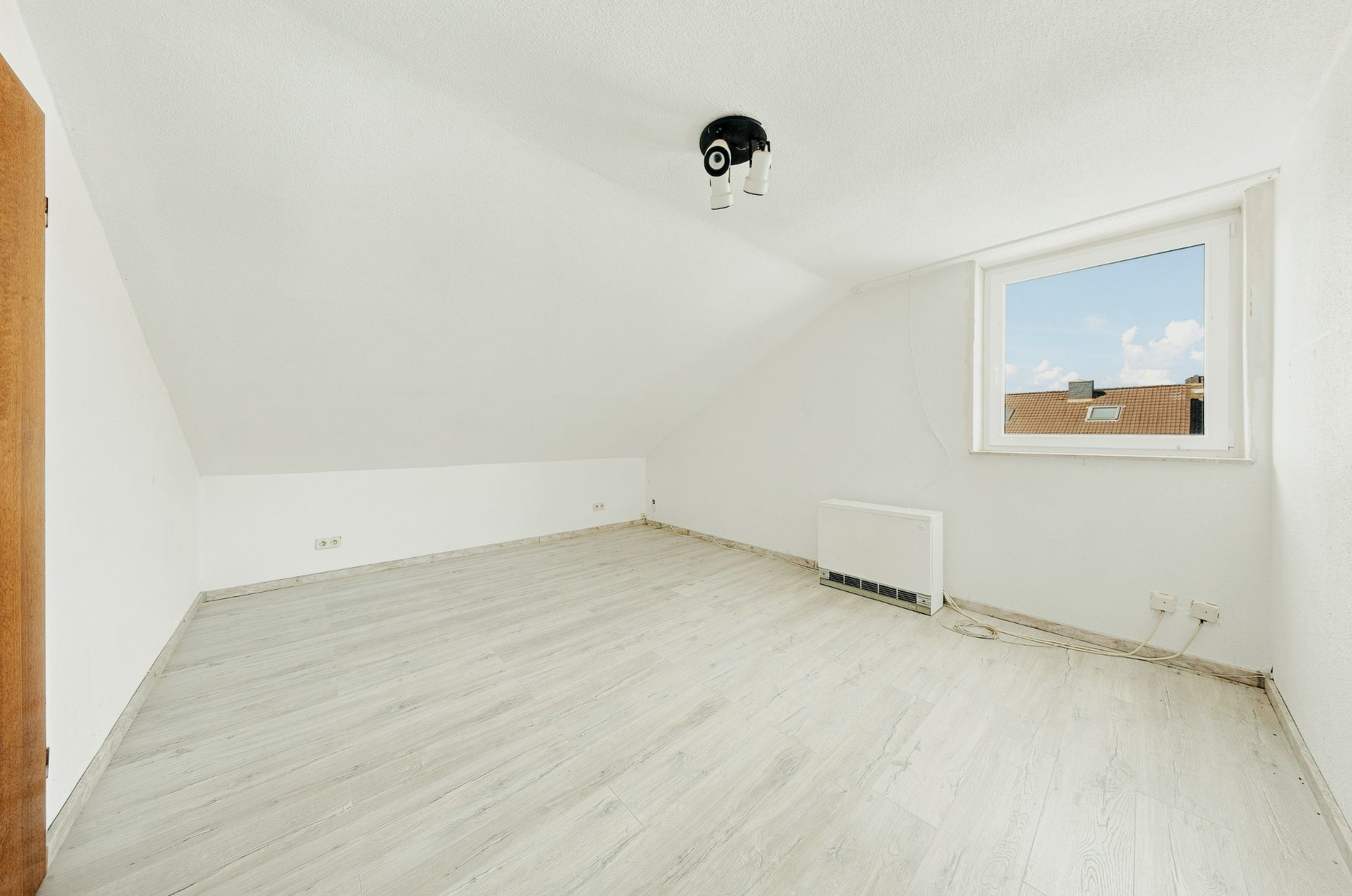 Leerer, heller Dachraum mit Holzboden, weißer Wand, Fenster und Heizkörper.