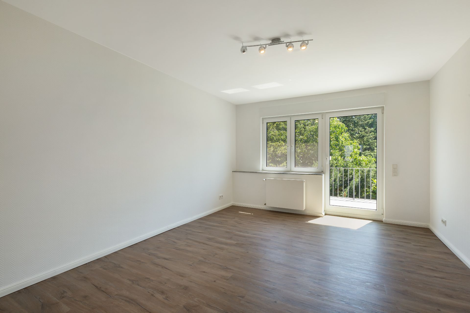 Helles, leeres Zimmer mit Holzfußboden, großem Fenster und Tür zu einem kleinen Balkon.
