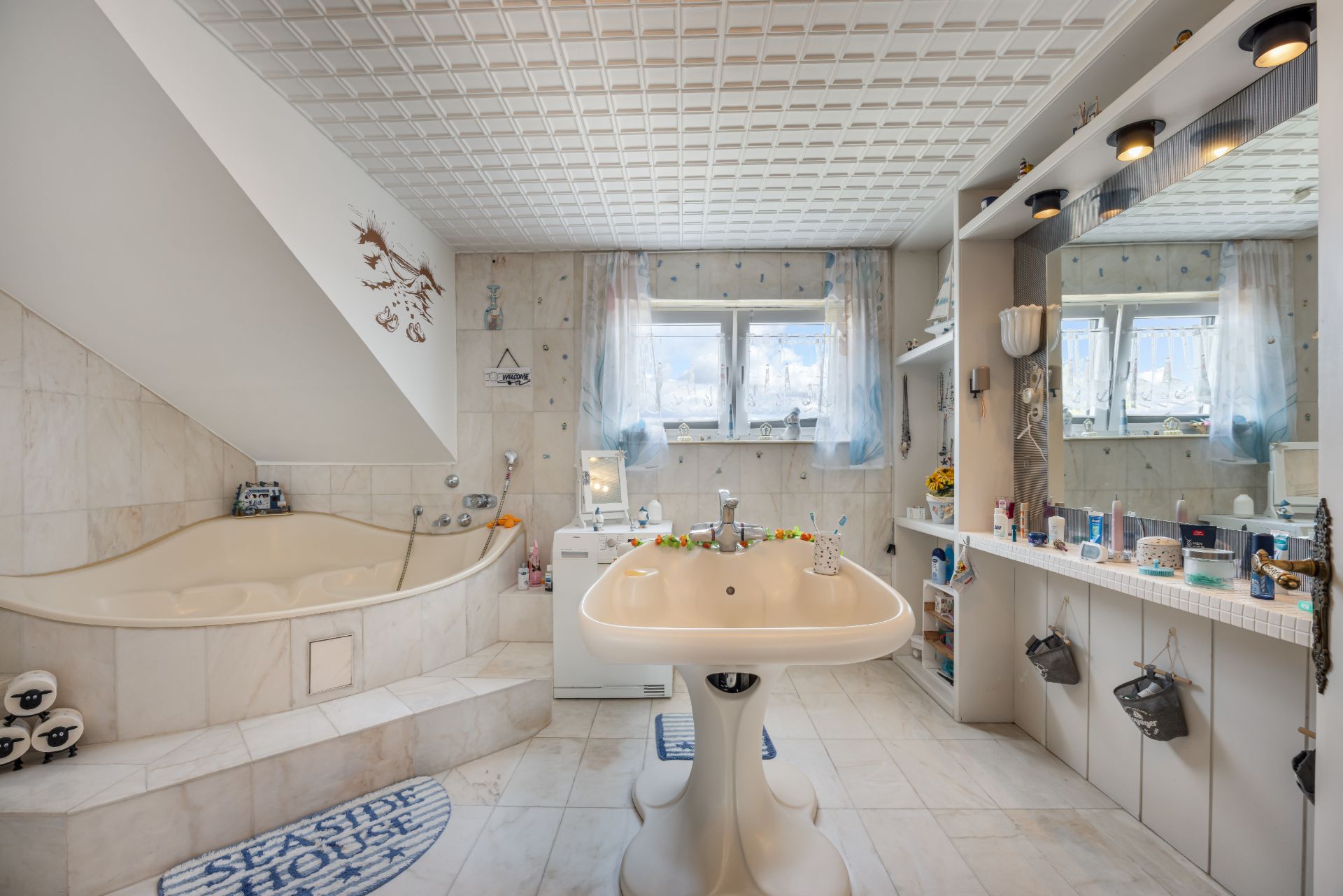 Helles Badezimmer mit Eckbadewanne, Waschbecken und großem Spiegel unter zwei Fenstern.