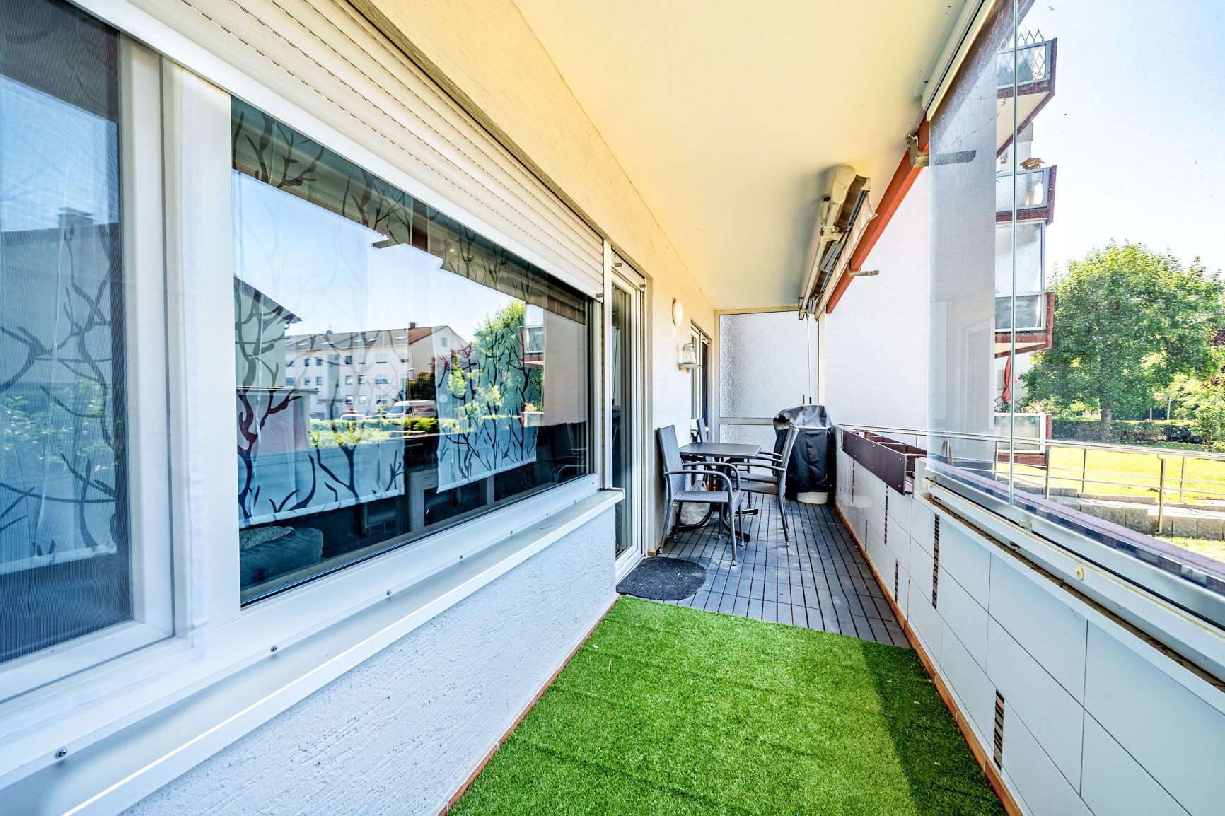 Langer Balkon mit Kunstrasen, Sitzmöbeln und Blick auf Wohngebäude und Bäume.