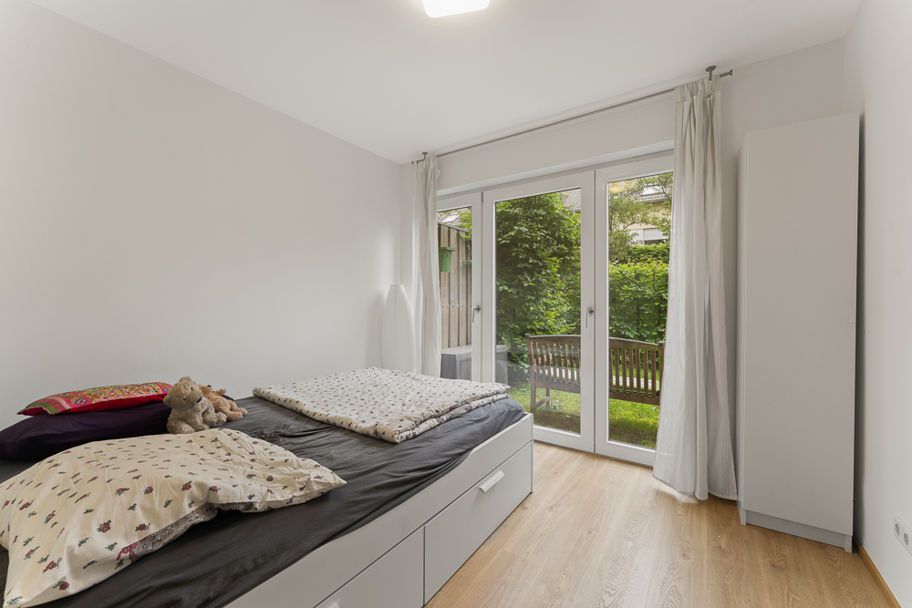 Helles Schlafzimmer mit Bett, Kleiderschrank und Glas­türen zum Garten mit Bank.