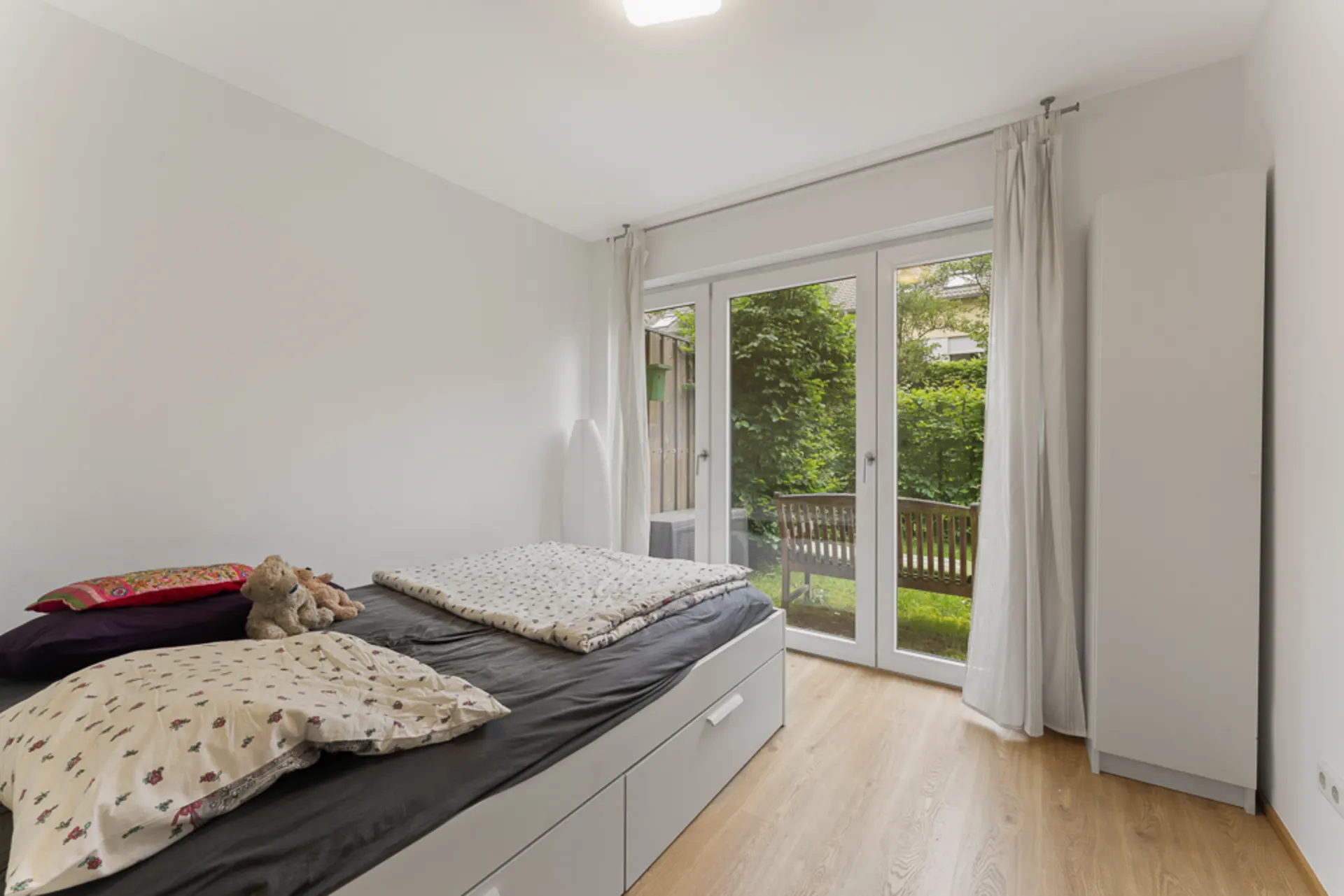 Helles Schlafzimmer mit Bett, Kleiderschrank und Glastüren zum Garten mit Bank.