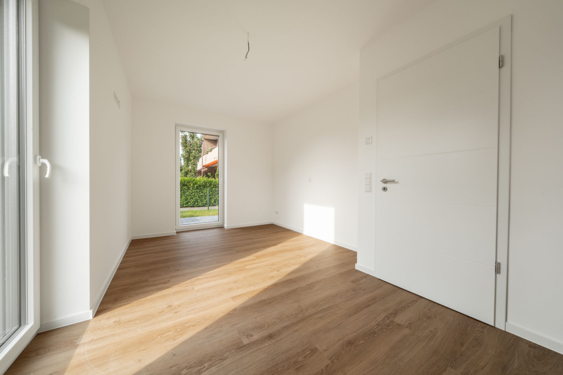 Heller Raum mit Holzfußboden, weißen Wänden und Tür, großes Fenster zur Terrasse.