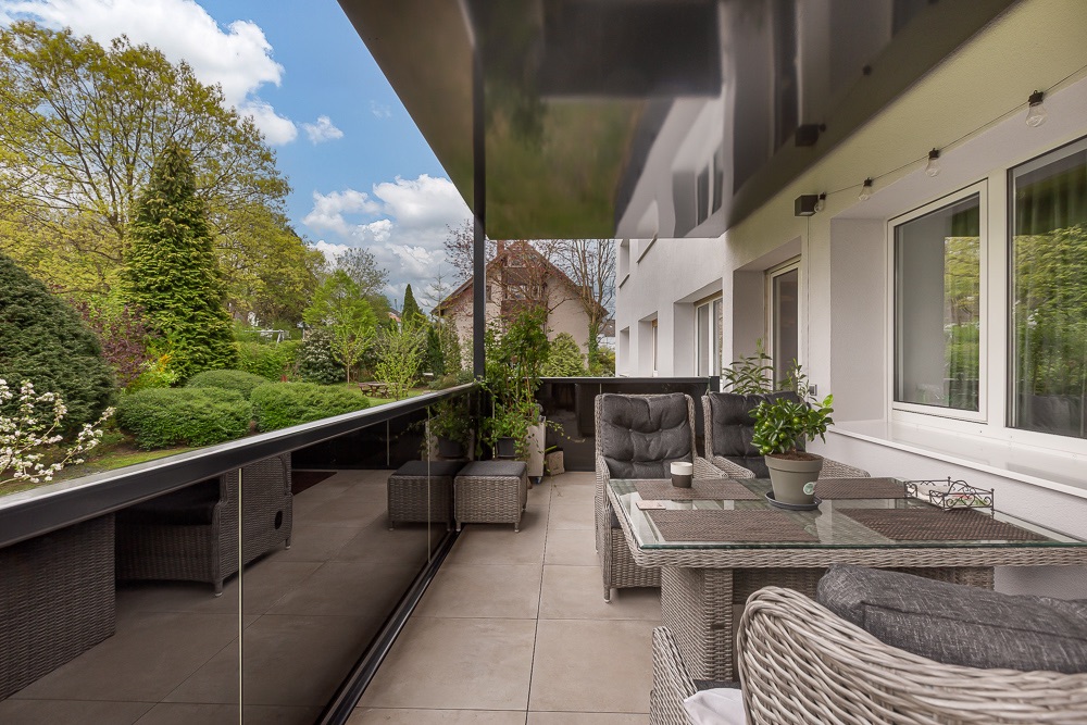 Moderne Terrasse mit Sitzmöbeln, Glastisch und Pflanzen, Blick auf Garten und Bäume.