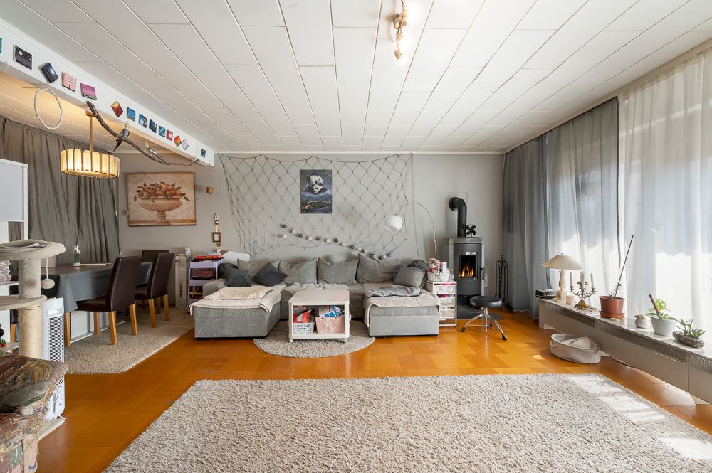 Wohnzimmer mit grauem Sofa, Teppich, Esstisch, Kaminofen und großen Fenstern mit Vorhängen.
