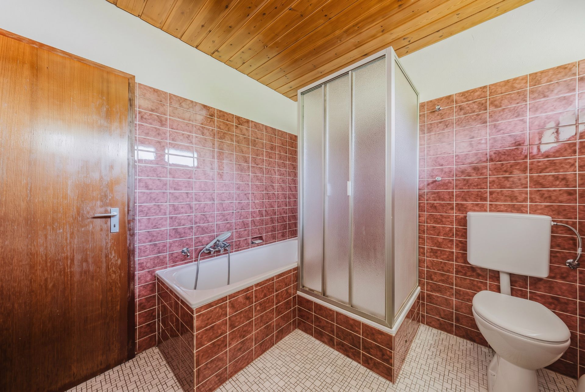 Badezimmer mit braunen Fliesen, Badewanne, Dusche und Toilette, Holztür und Holzdecke.
