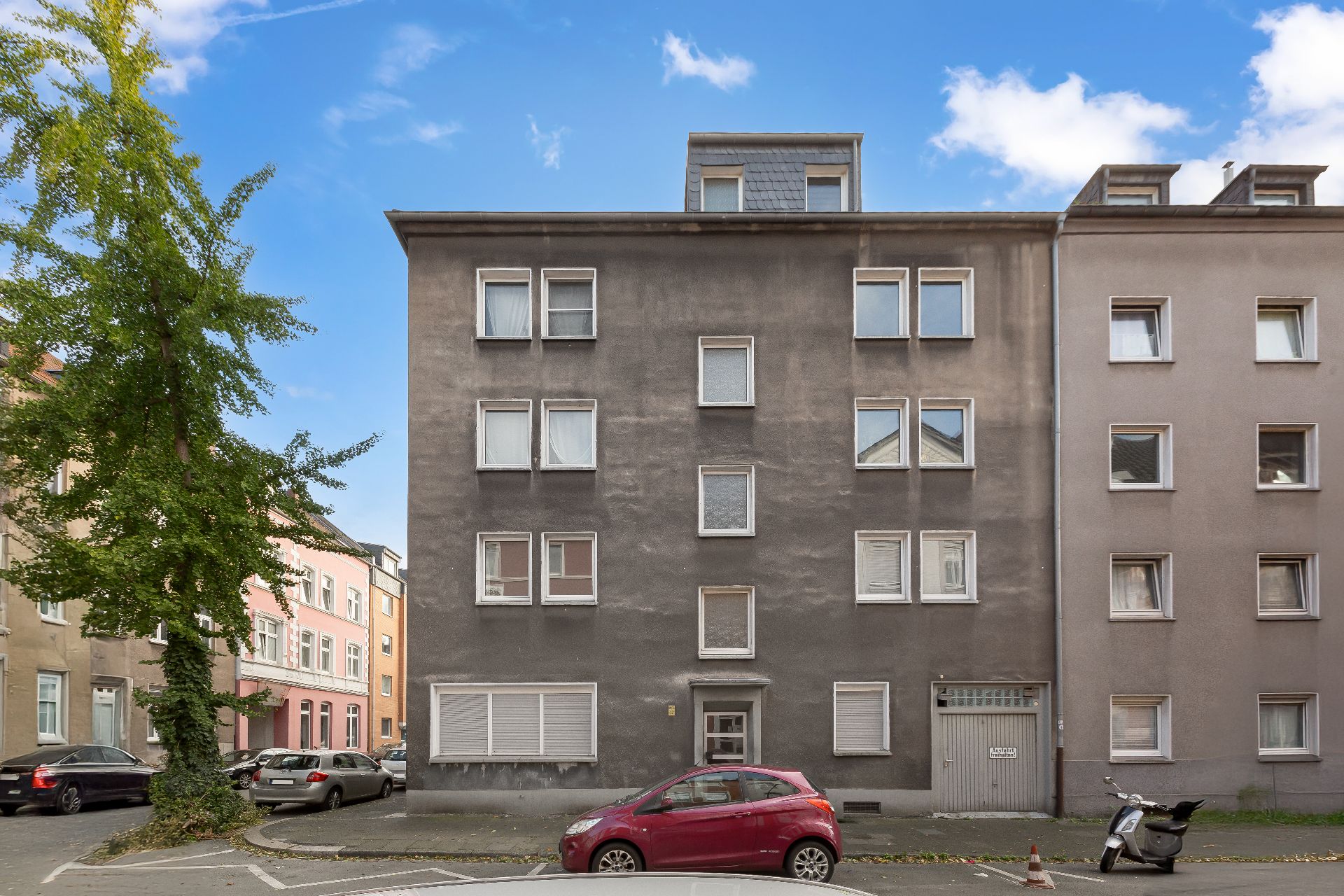 Wohnhaus mit grauer Fassade, roten Auto und Roller davor, Bäume und andere Gebäude nebenan.
