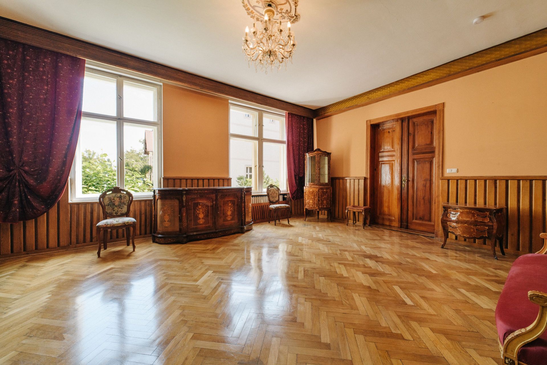 Helles Wohnzimmer mit Parkettboden, Kronleuchter, großen Fenstern und antiken Holzmöbeln.