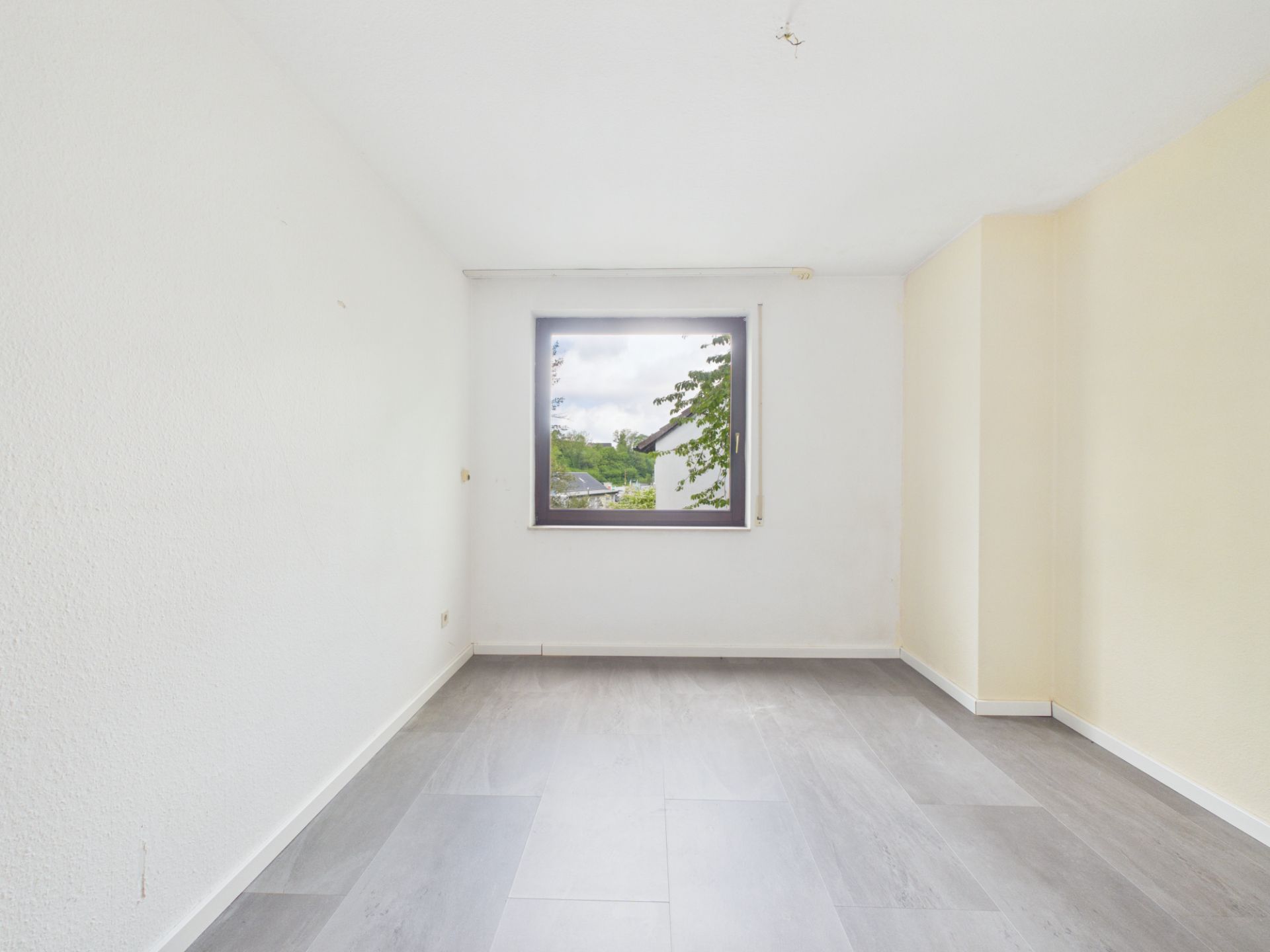 Leeres Zimmer mit grauem Boden und großem Fenster, das auf Bäume und Himmel blickt.