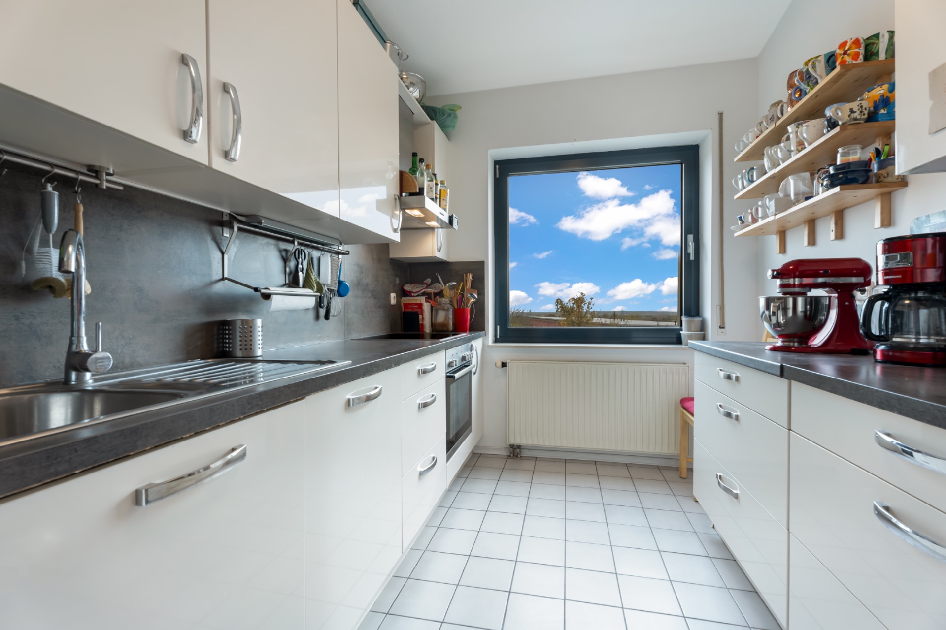 Moderne Küche mit weißen Schränken, Geräten, Regalen und großem Fenster mit Ausblick.