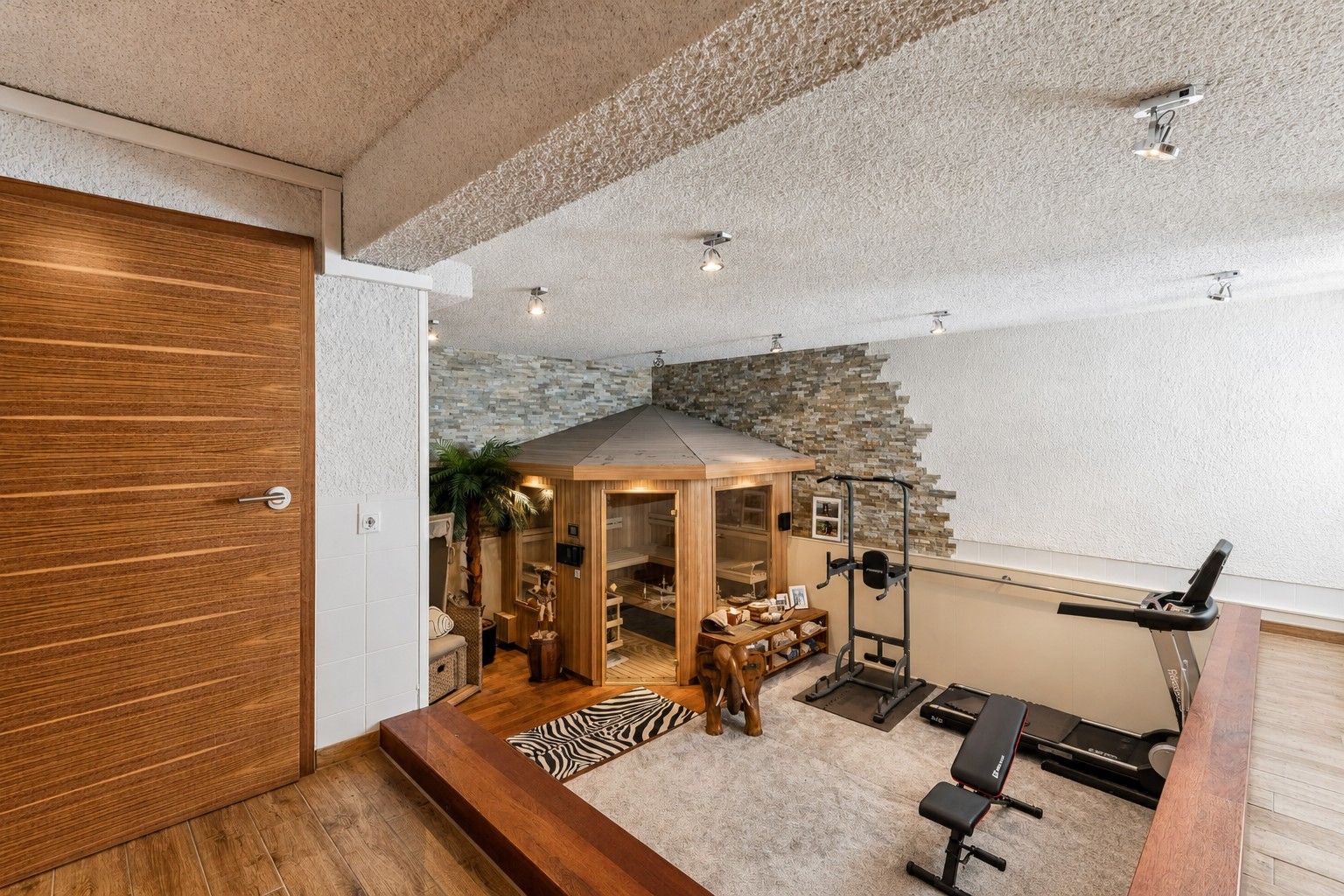 Wohnraum mit Holzsauna, Fitnessgeräten und Dekoration, heller Boden und Steinwand.