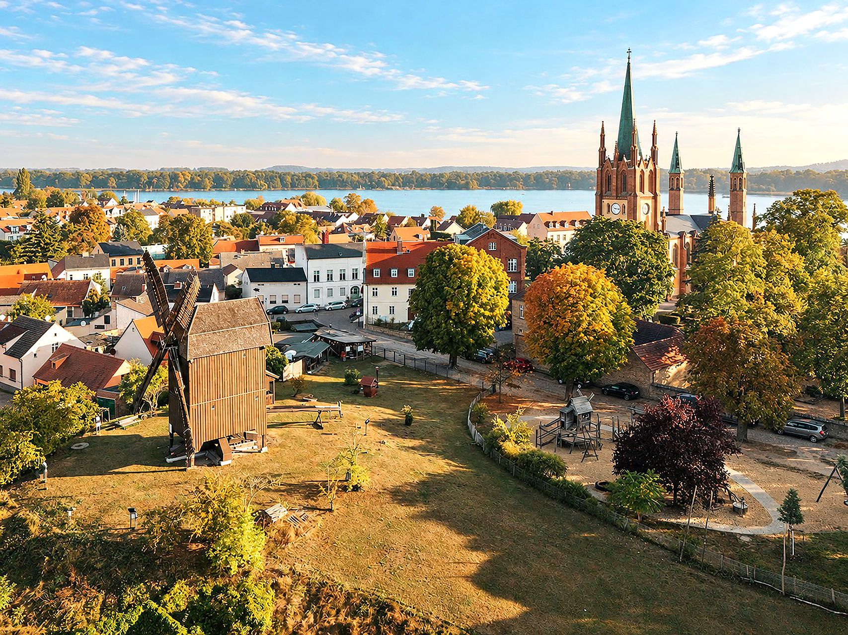 Stadtansicht mit einer Windmühle, Bäumen, Häusern und einer großen Kirche am Wasser.