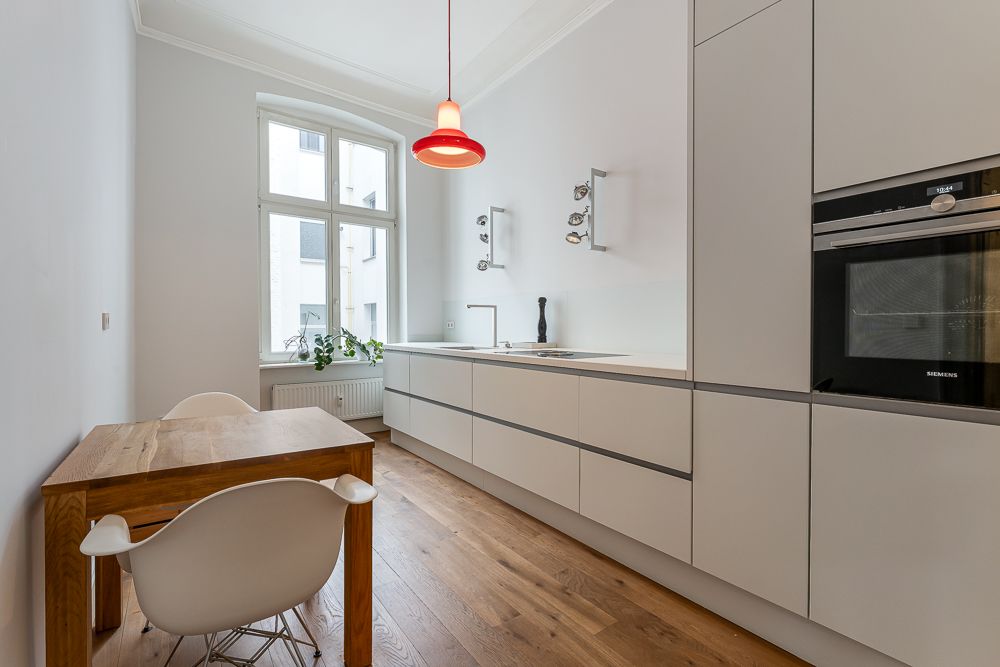 Helle, moderne Küche mit Holzboden, Einbauküche, Tisch und Stuhl, großem Fenster und roter Hängelampe.