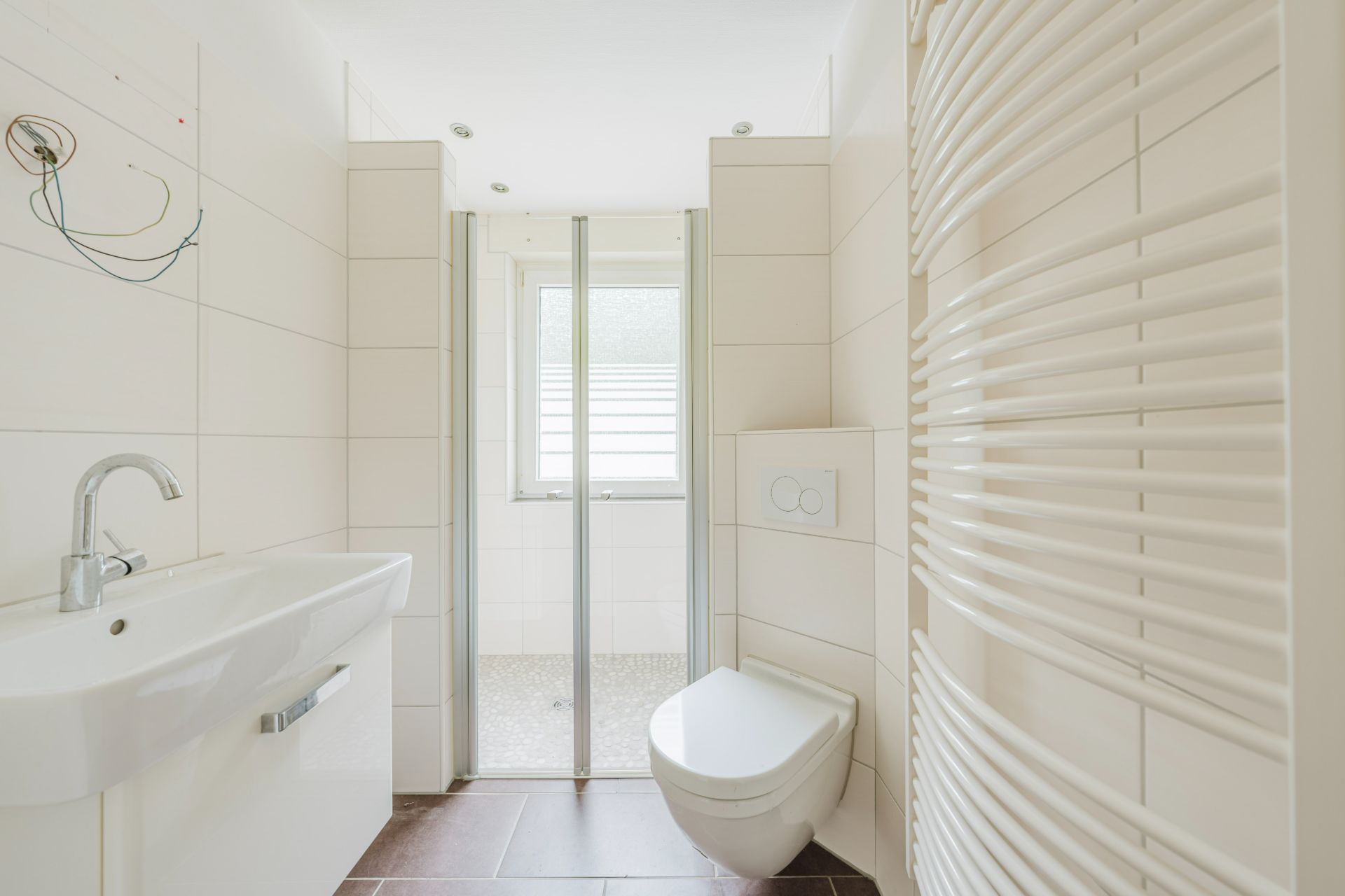 Helles Badezimmer mit Waschbecken, Toilette, Handtuchheizkörper und Dusche mit Glasabtrennung.