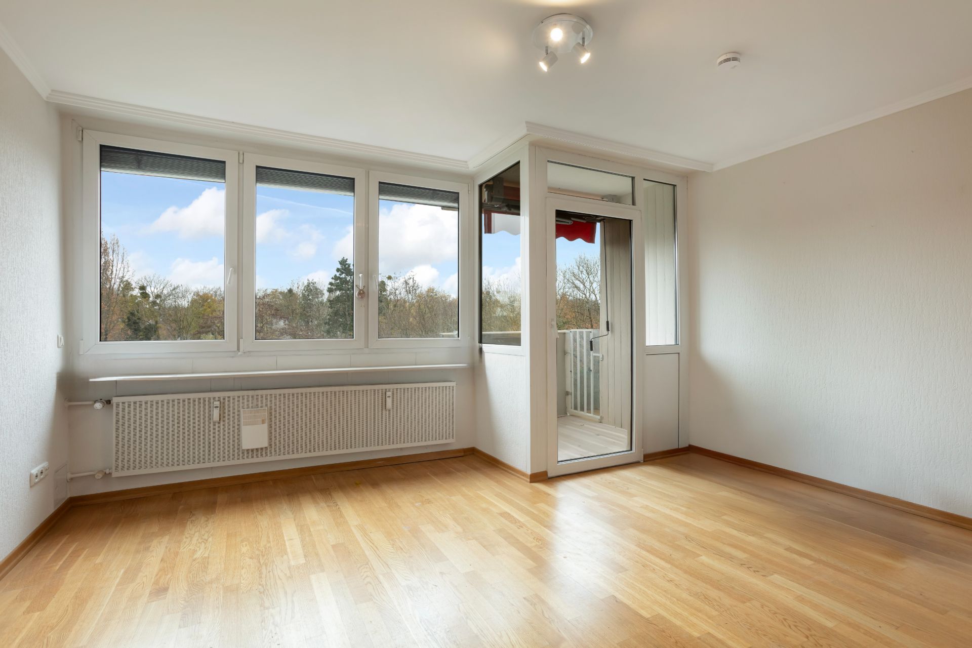 Helles Zimmer mit Holzfußboden, großen Fenstern und Zugang zu einem verglasten Balkon.
