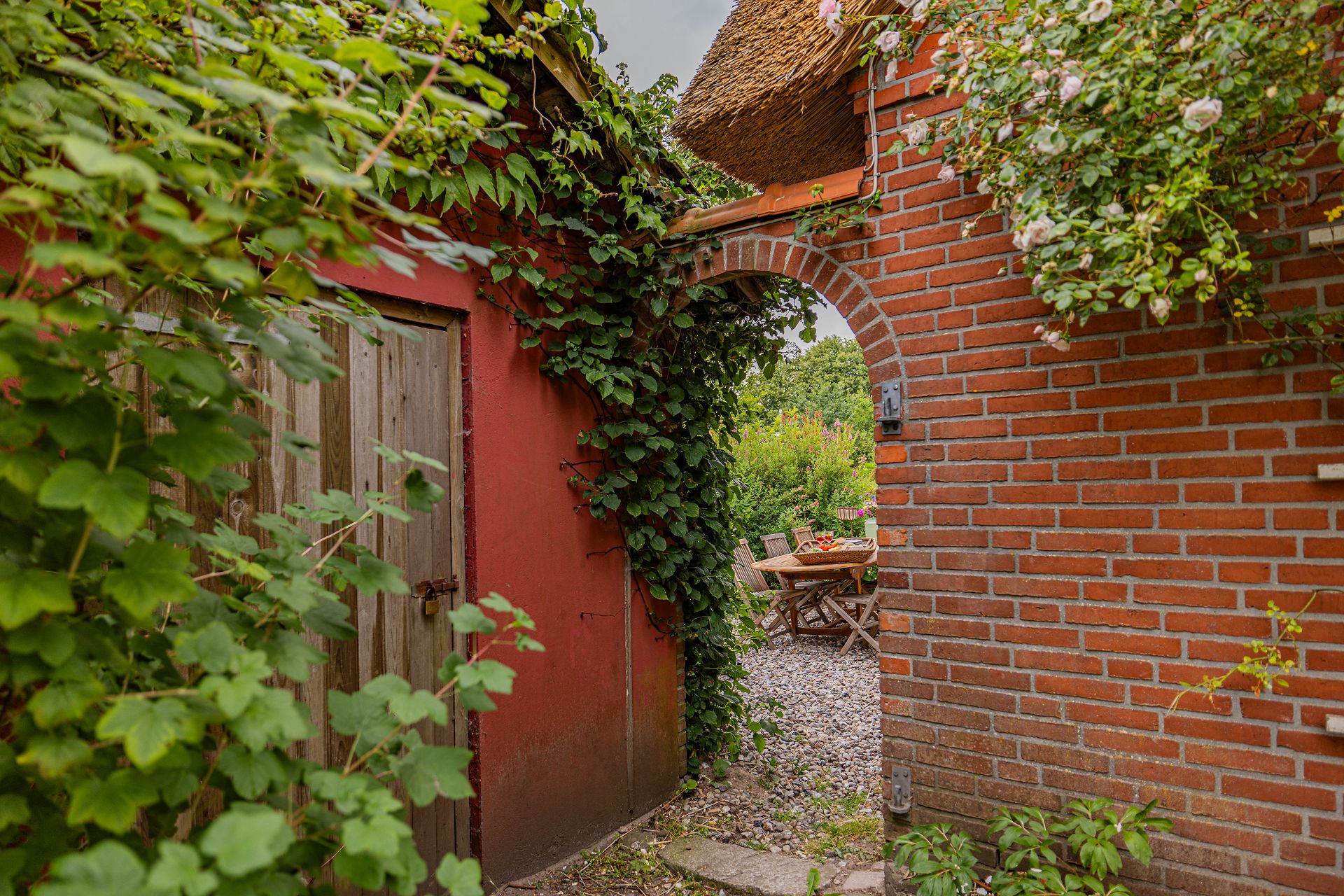 Garten mit roten Backsteinmauern, Holztür, viel Grün und Sitzbereich mit Holztisch und Stühlen.