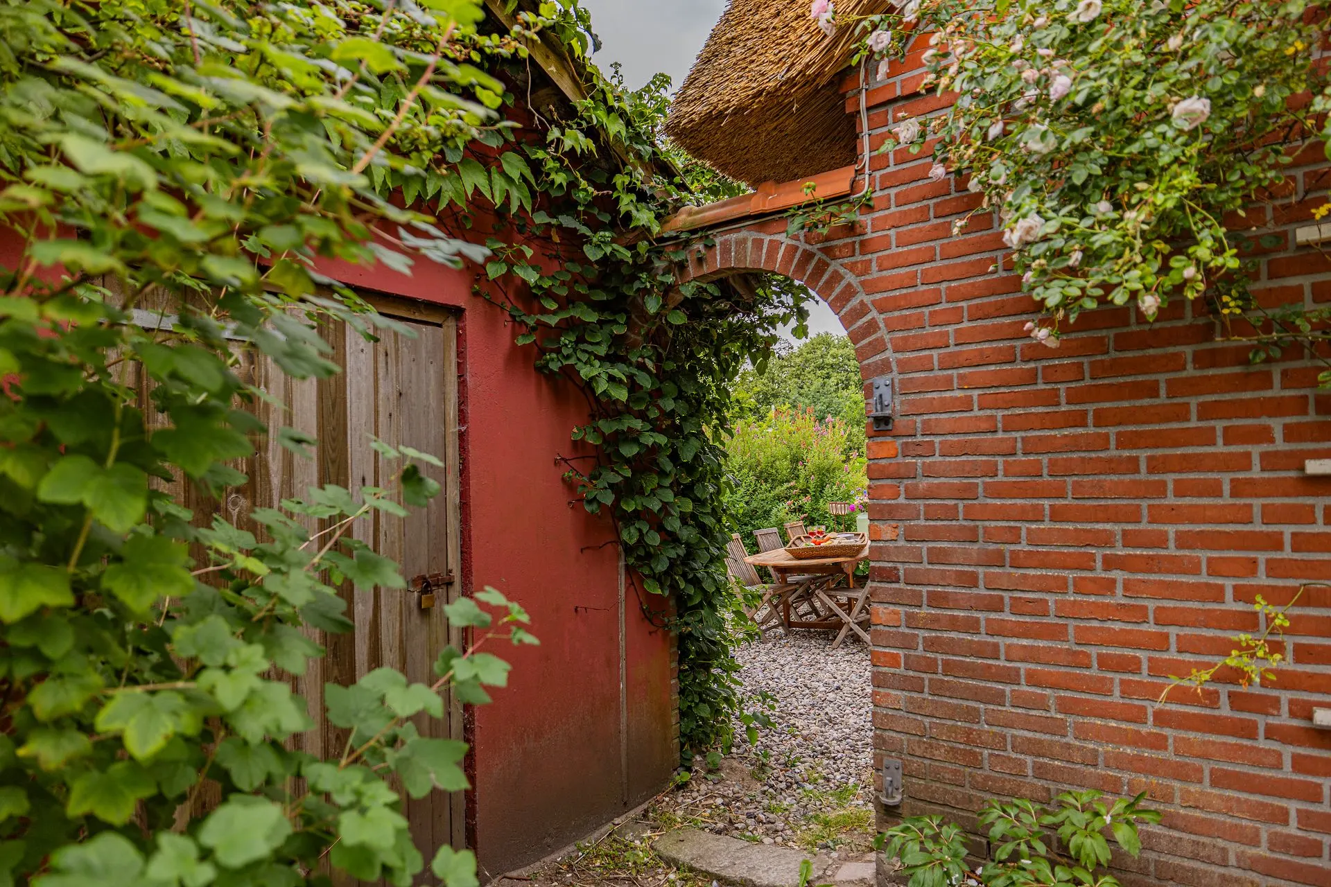 Garten mit roten Backsteinmauern, Holztür, viel Grün und Sitzbereich mit Holztisch und Stühlen.