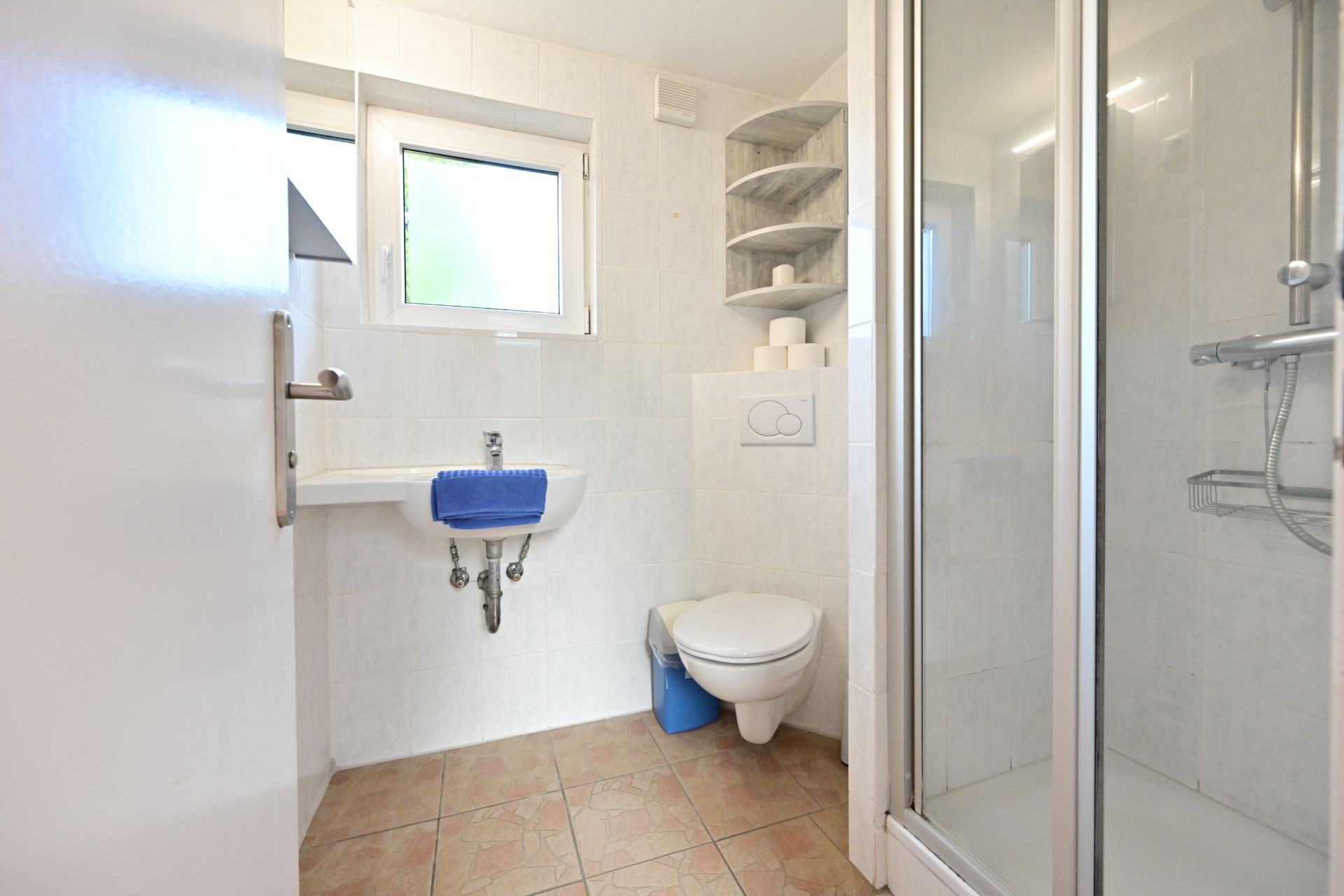 Helles Badezimmer mit Dusche, WC, Waschbecken, Fenster und Regal mit Toilettenpapierrollen.