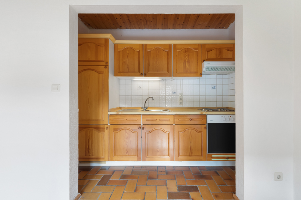 Kleine Küche mit Holzschränken, Herd, Spüle und weißen Fliesen an der Wand.