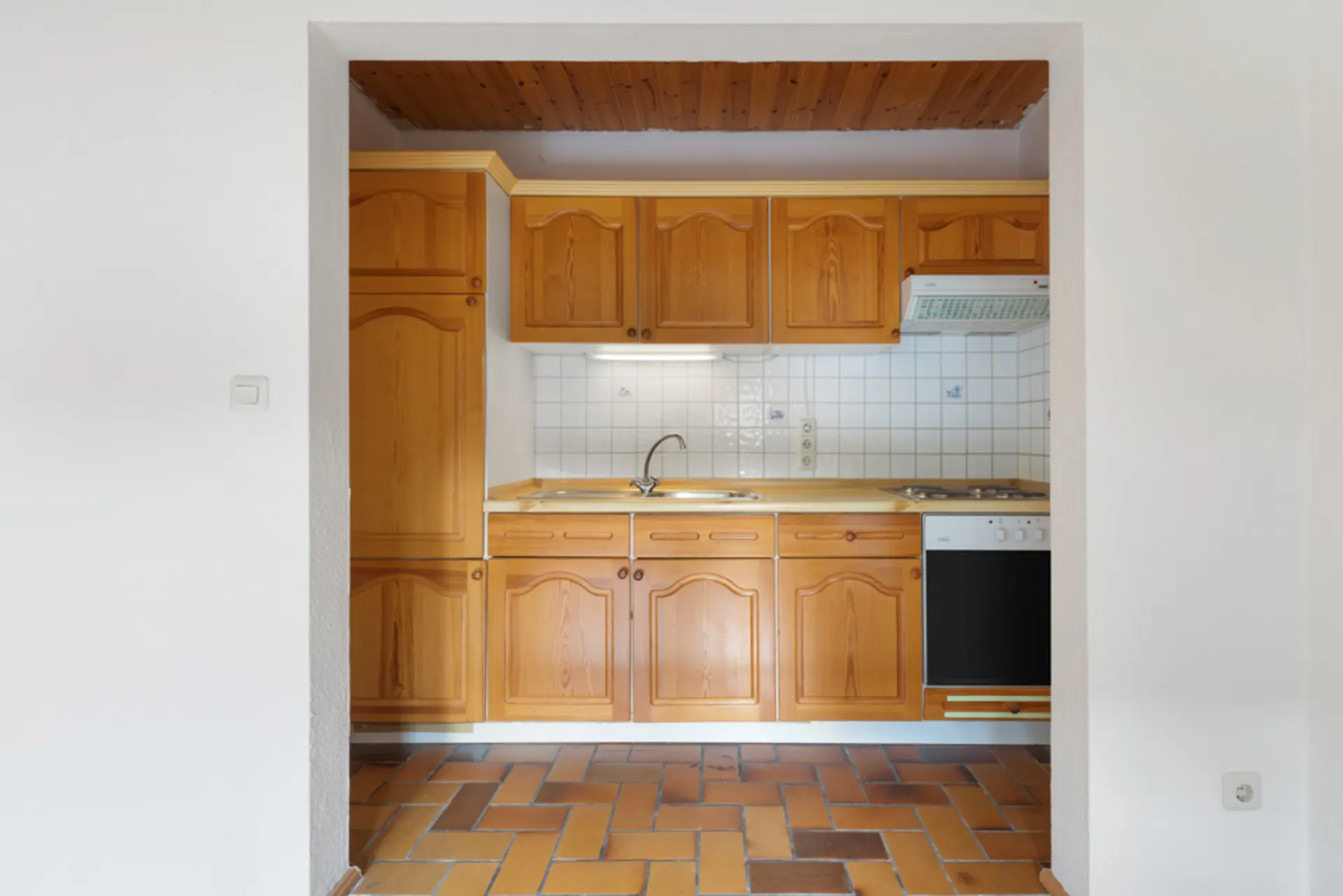 Kleine Küche mit Holzschränken, Herd, Spüle und weißen Fliesen an der Wand.