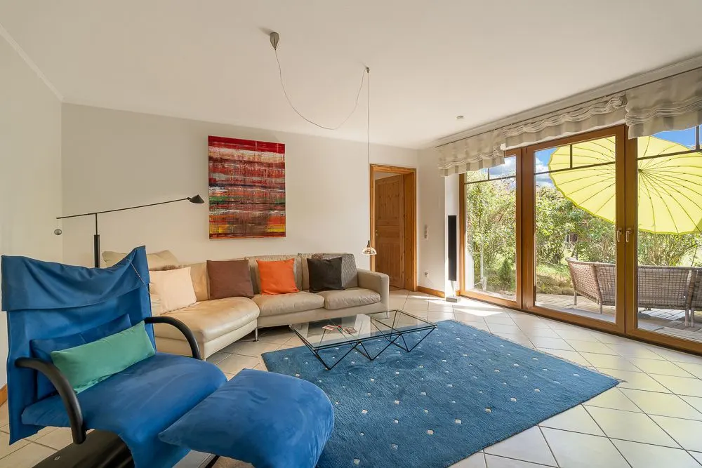 Helles Wohnzimmer mit Sofa, blauem Sessel, Glastisch und großem Fenster zum Garten.