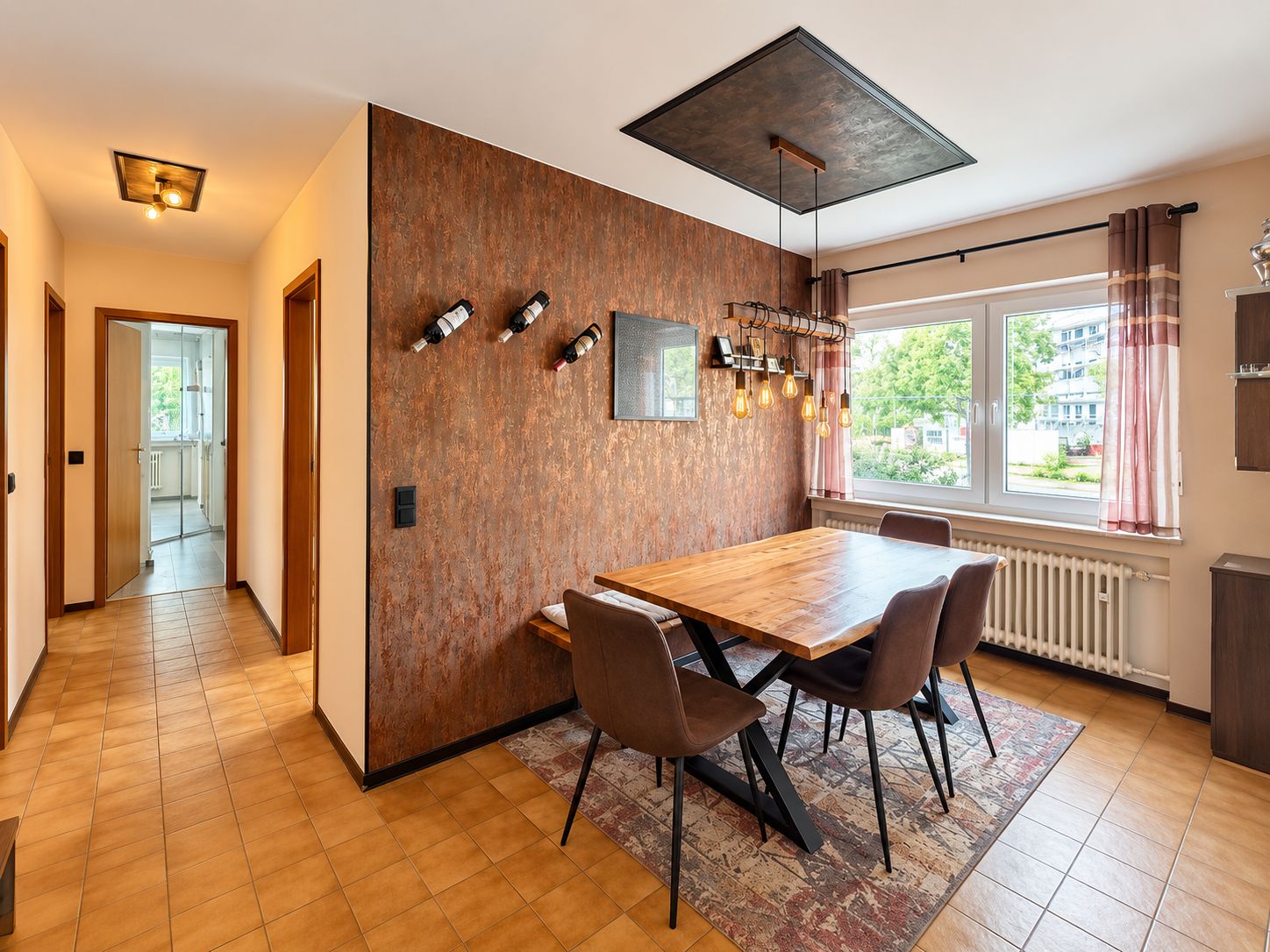 Essbereich mit Holztisch, vier Stühlen, Fenster und moderner Beleuchtung an brauner Wand.