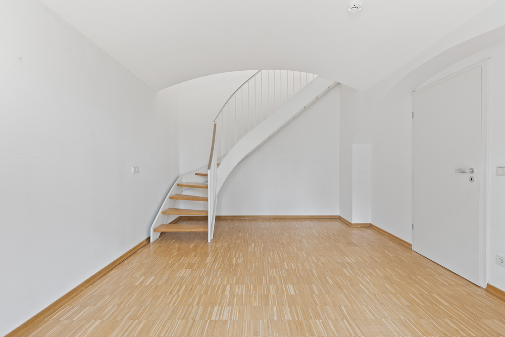 Heller Raum mit Holzfußboden, weißer Wand und einer offenen Holztreppe mit weißem Geländer.