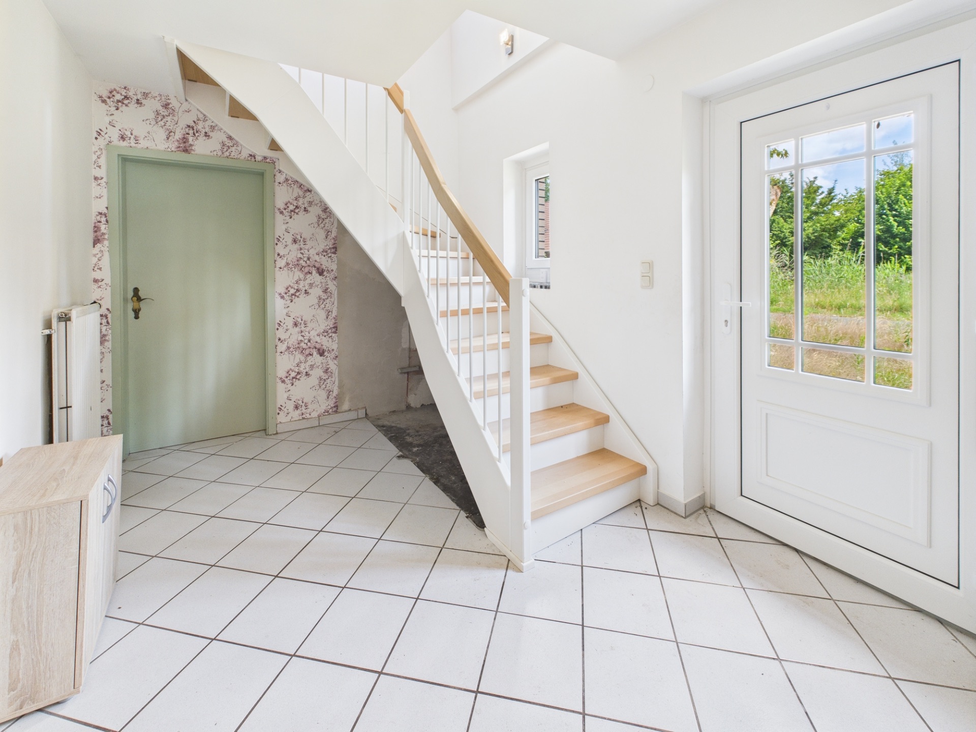Heller Flur mit weißer Treppe, grüner Tür, Fliesenboden und Glastür zum Garten.